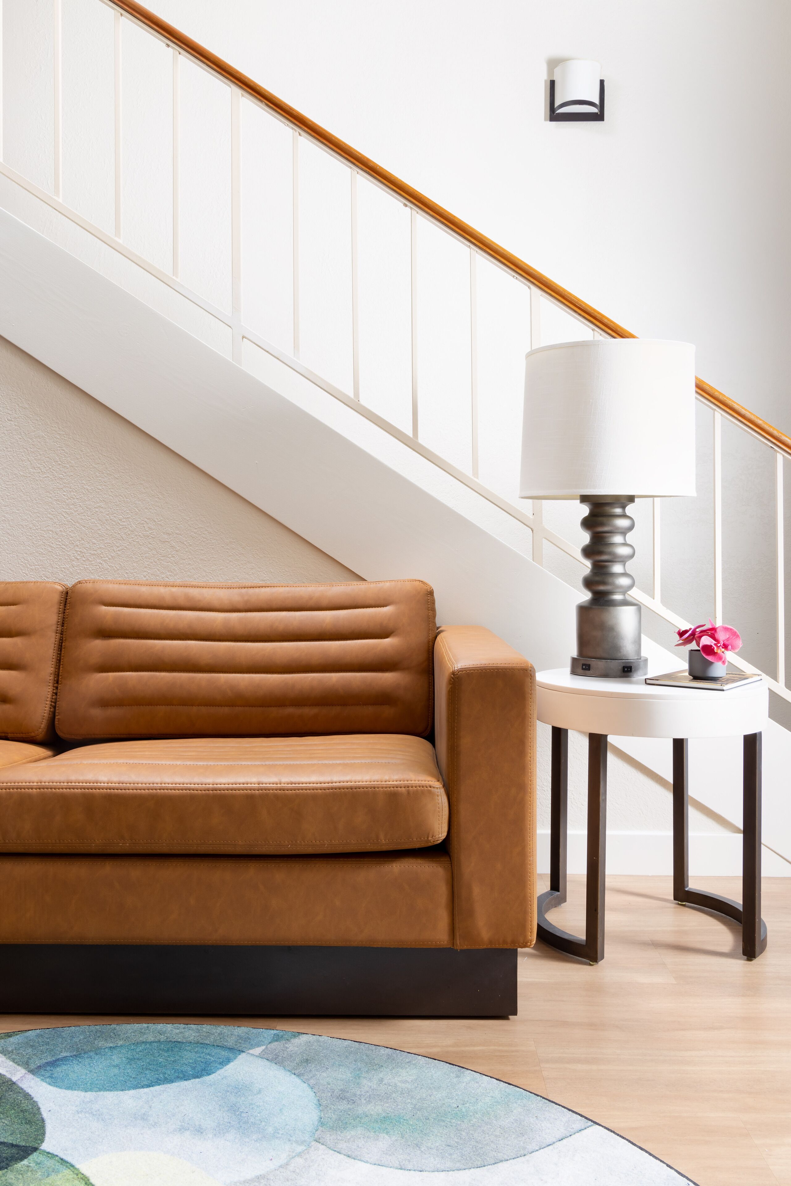 Ein braunes Ledersofa, ein runder Beistelltisch mit einer Lampe und ein farbenfroher Teppich in einem Zimmer mit einer Holztreppe.