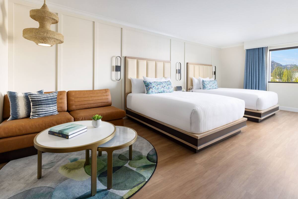 A hotel room with a sofa, two beds, a round coffee table, and a view of the outdoors.
