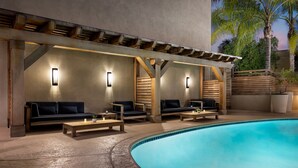 Zona de piscina con sillones reclinables de madera y un banco integrado, bajo una pérgola con luces fijadas a la pared.