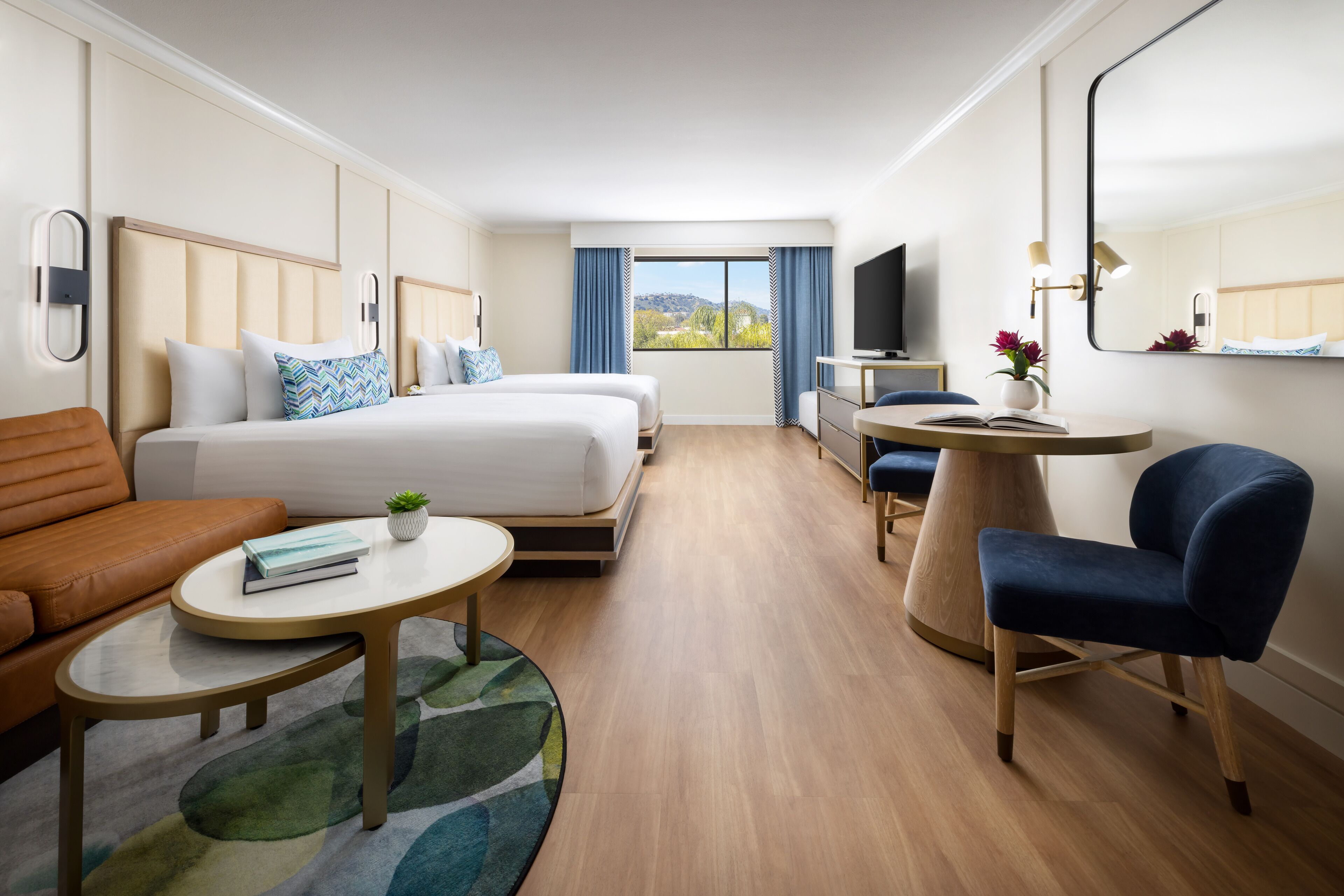 Quarto de hotel com duas camas, um sofá, uma pequena mesa, uma cadeira, uma televisão e uma janela com vista.