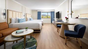 Quarto de hotel com duas camas, um sofá, uma pequena mesa, uma cadeira, uma televisão e uma janela com vista.
