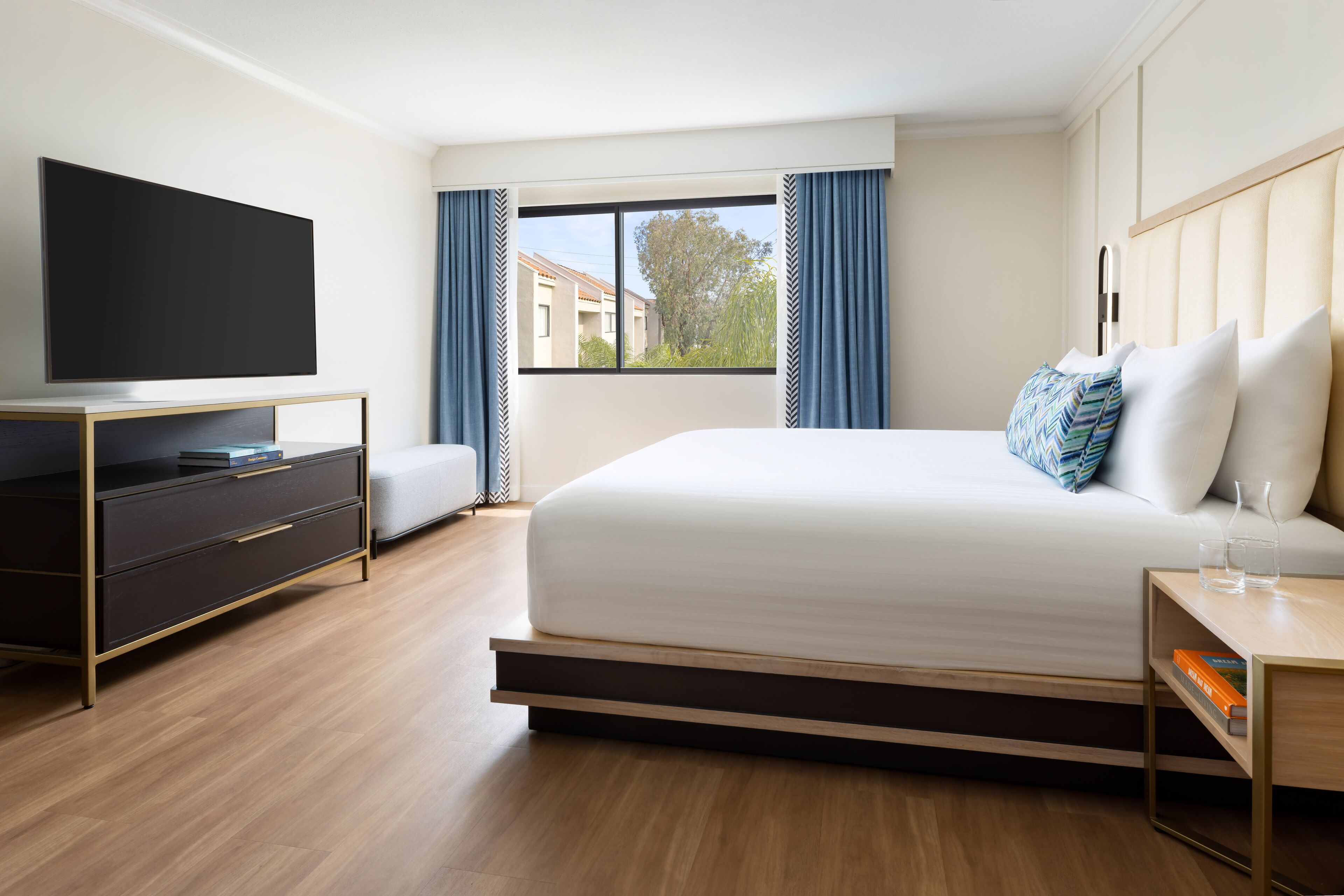 A modern hotel room with a large bed, a flat-screen TV, a wooden desk, and a window with curtains.