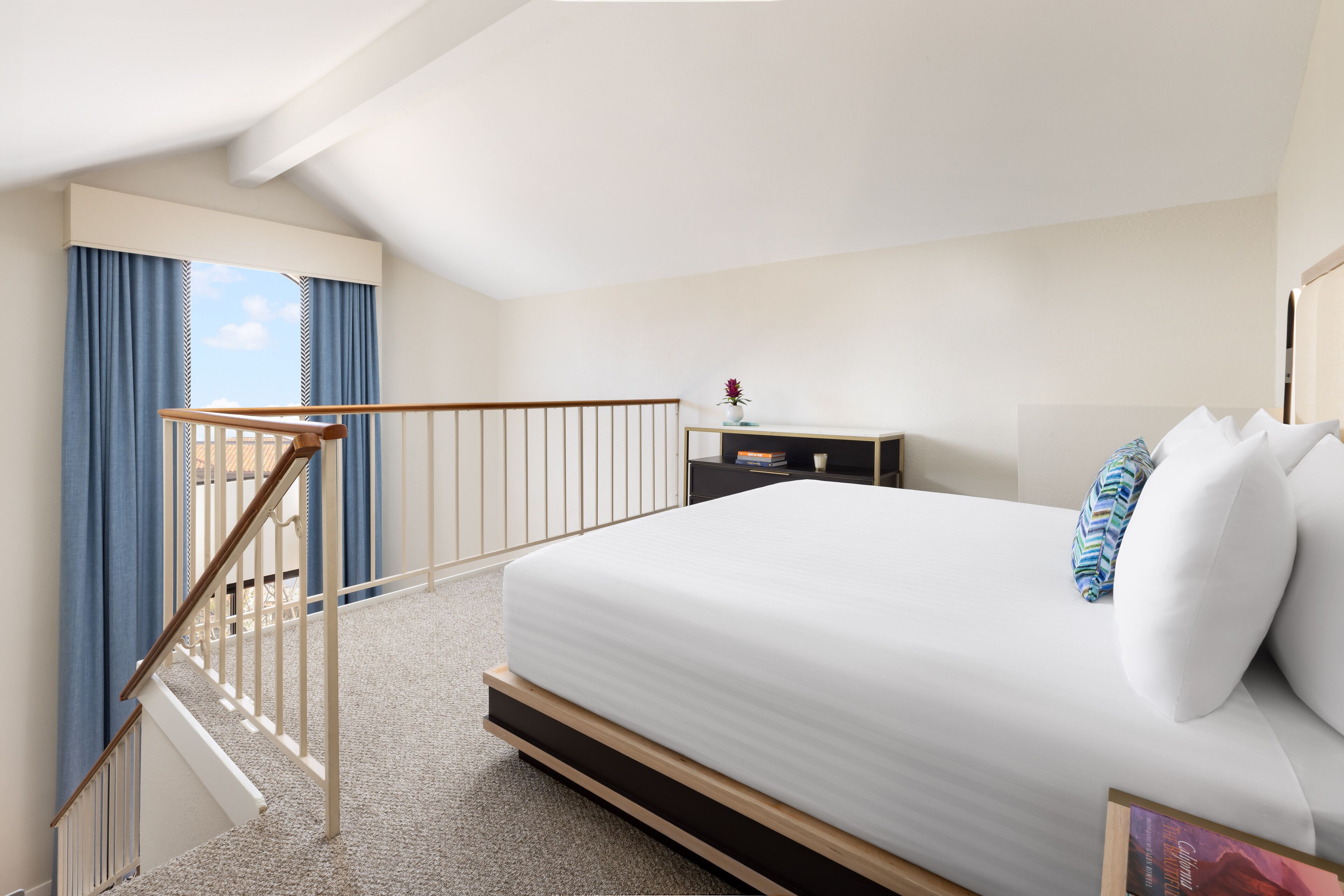 A hotel room with a bed, a nightstand, a dresser, a window with curtains, and a staircase.