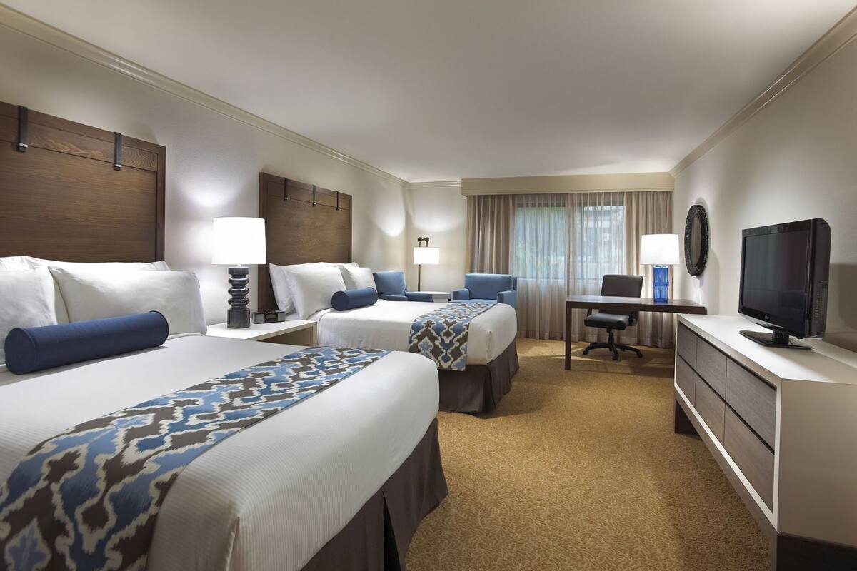 A hotel room with two beds, a desk, a television, and a large window with curtains.