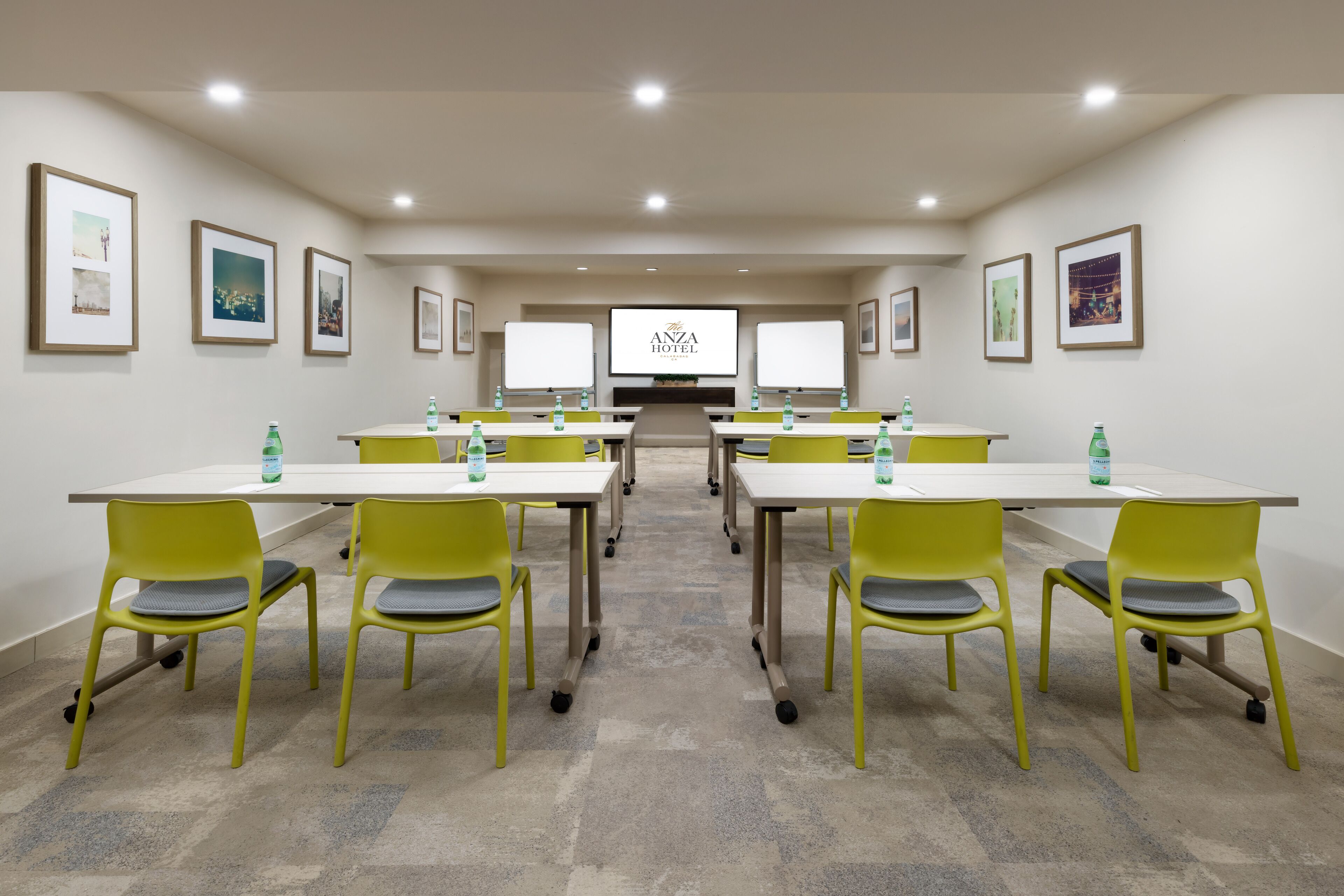 A conference room with a large screen displaying "ANZA HOTEL" at the front. There are six tables with yellow chairs, each table has a bottle of water. The room has framed pictures on the walls and a carpeted floor.