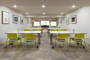 A conference room with a large screen displaying "ANZA HOTEL" at the front. There are six tables with yellow chairs, each table has a bottle of water. The room has framed pictures on the walls and a carpeted floor.