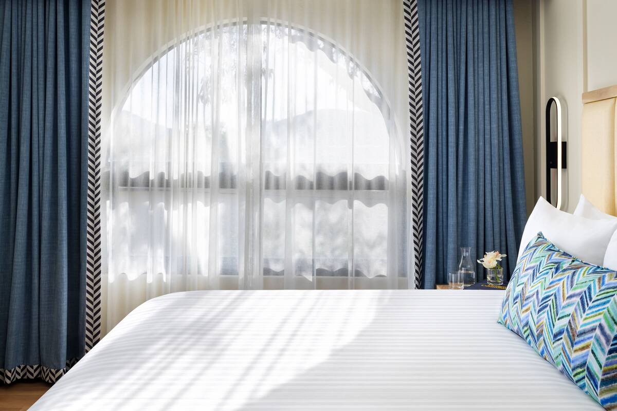 A hotel room with a large bed, a mirror, and a view of the outdoors through sheer curtains.