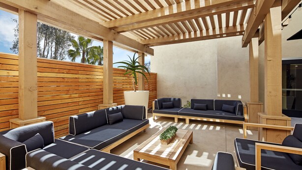 Un patio exterior con mobiliario de madera, incluyendo sofás y una mesa de centro, bajo una pérgola de madera.