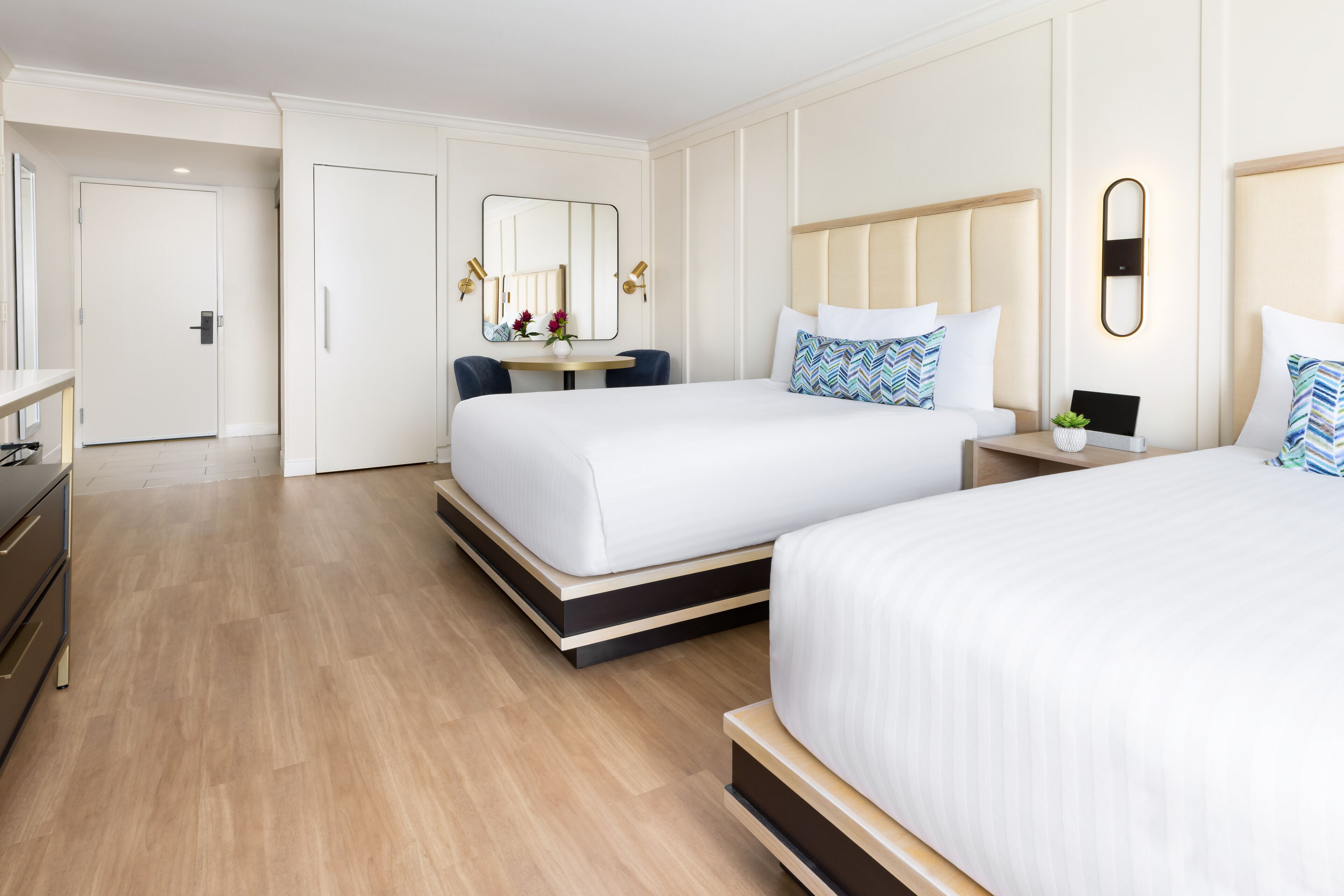 A modern hotel room with two beds, a small desk, a chair, and a large mirror.