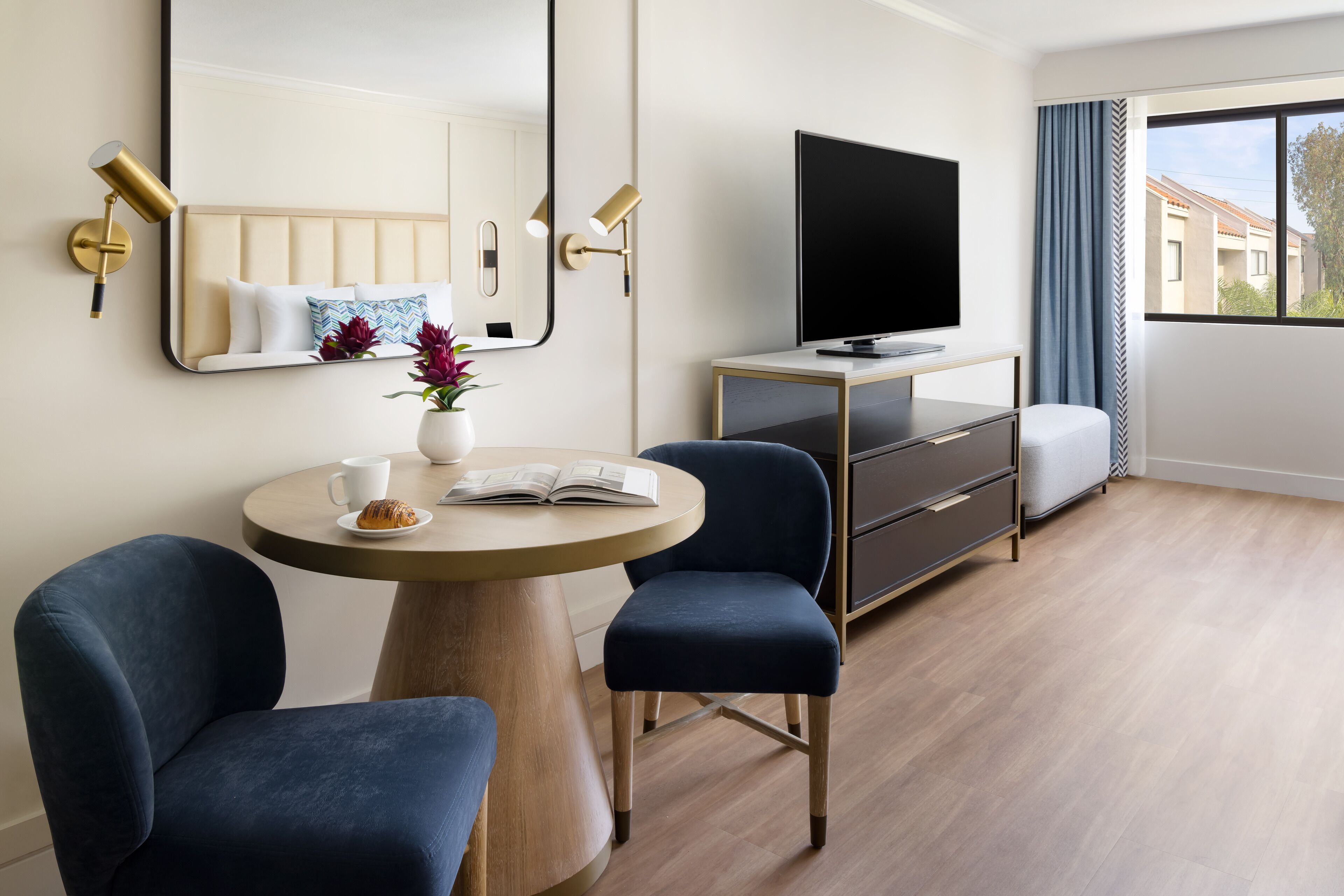 A modern hotel room with a round wooden table, two blue chairs, a TV on a wooden cabinet, a large mirror, and a window with a view of buildings.
