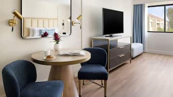 A modern hotel room with a round wooden table, two blue chairs, a TV on a wooden cabinet, a large mirror, and a window with a view of buildings.
