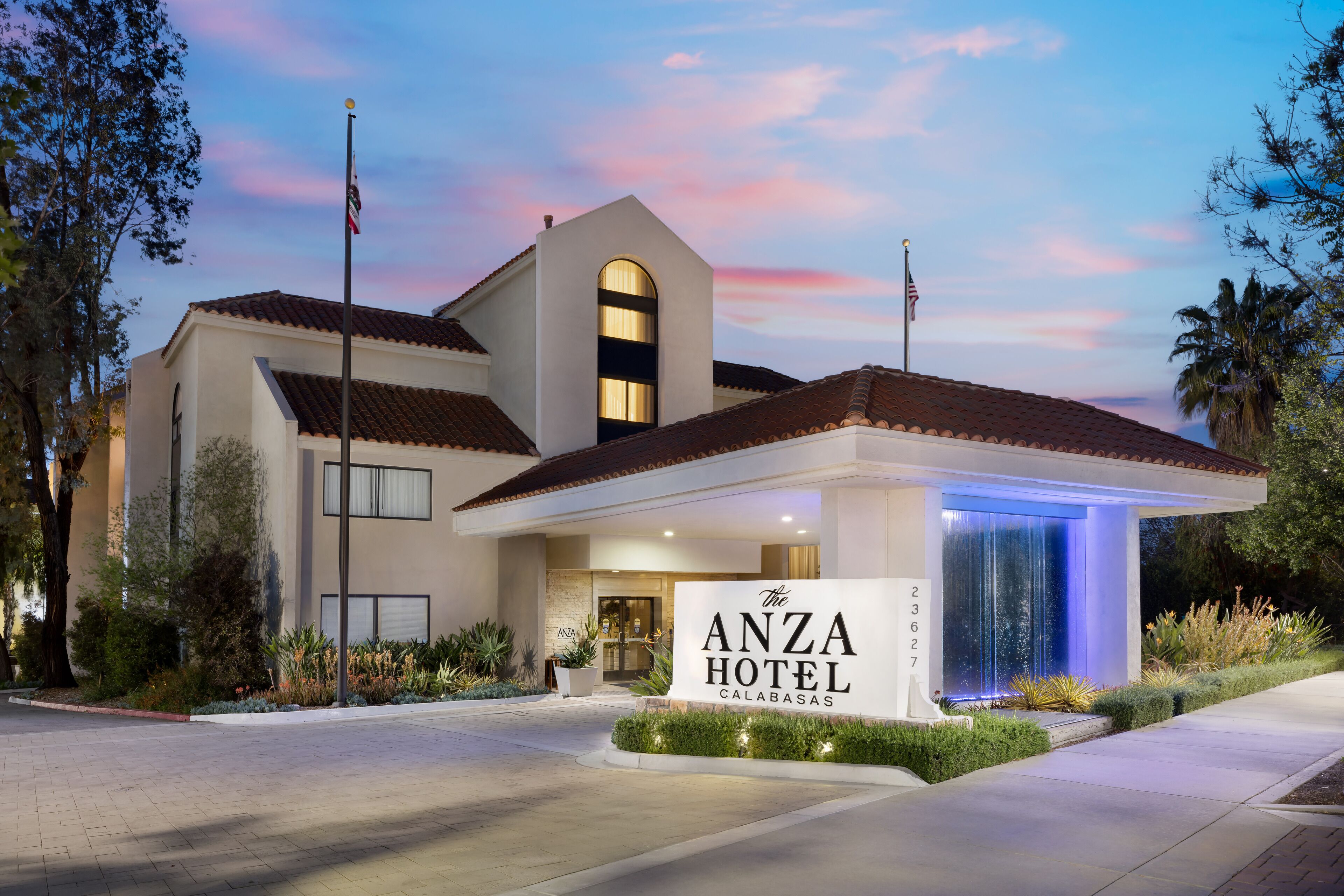 Exterior del Hotel Anza, con un estilo arquitectónico moderno que destaca una entrada imponente, un asta de banderas y jardines bien cuidados.
