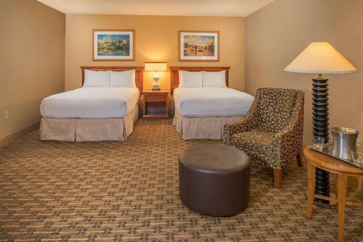 A hotel room with two beds, a patterned carpet, a patterned armchair, a round ottoman, a lamp, and a small table with a tray.