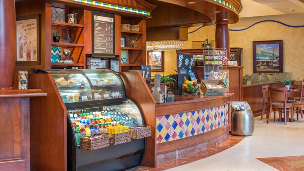 A deli counter with a glass display case filled with various food items, a menu board, and a decorative plant. - Hilton Seattle Airport & Conference Center (SeaTac)