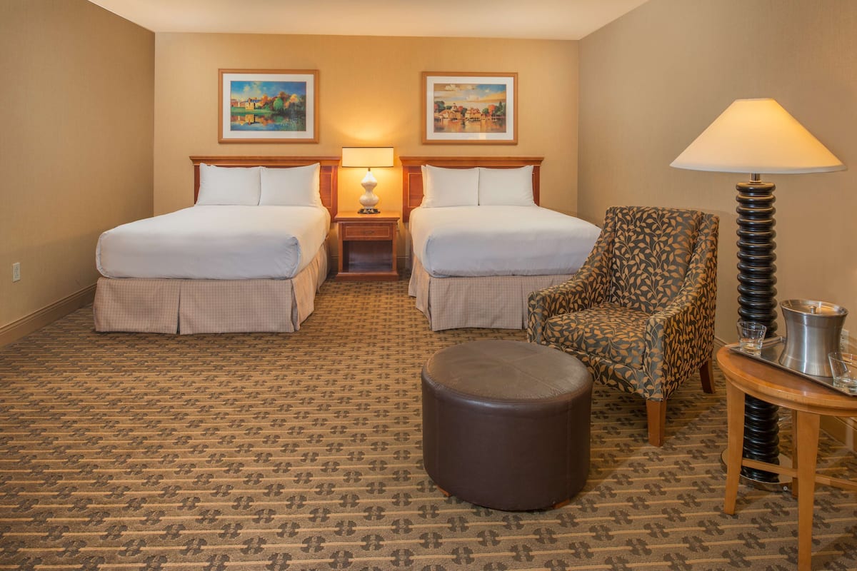 Une chambre d’hôtel avec deux lits, une moquette à motifs, un fauteuil à motifs, un pouf rond, une lampe et une petite table avec un plateau.