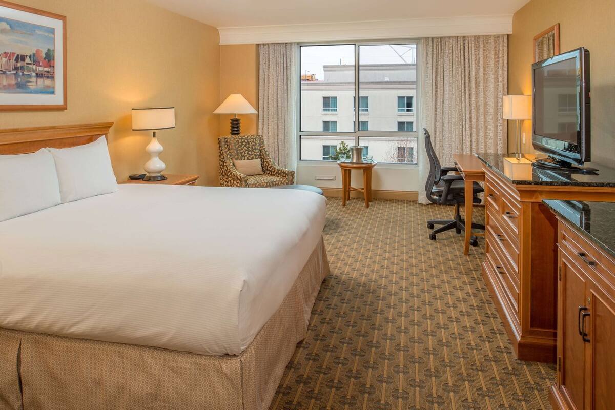 A hotel room with a large bed, a desk with a television, a chair, and a window with a view of buildings.
