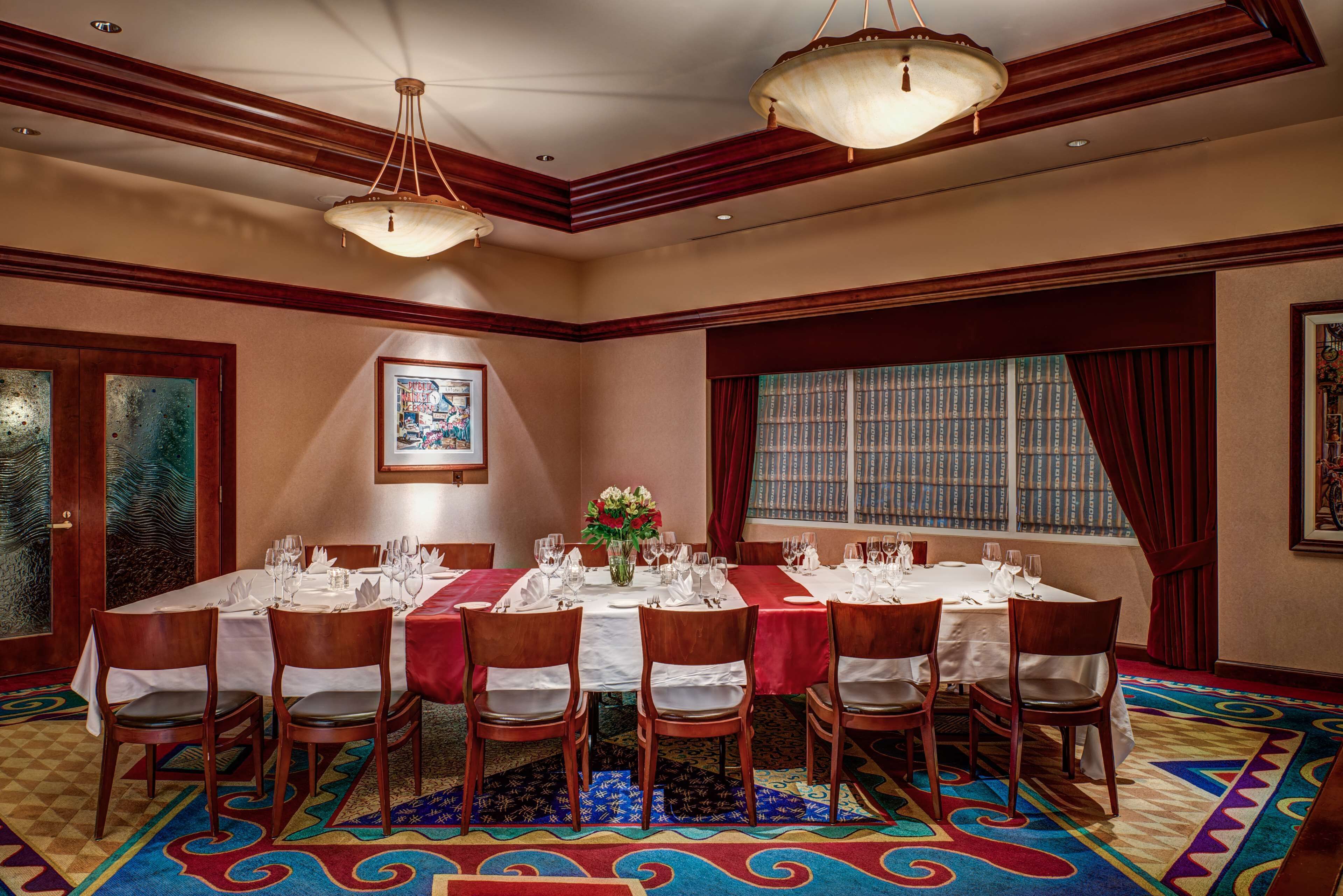 A dining room with a large table set for eight, surrounded by wooden chairs. There are two chandeliers, a framed picture on the wall, and a window with curtains.