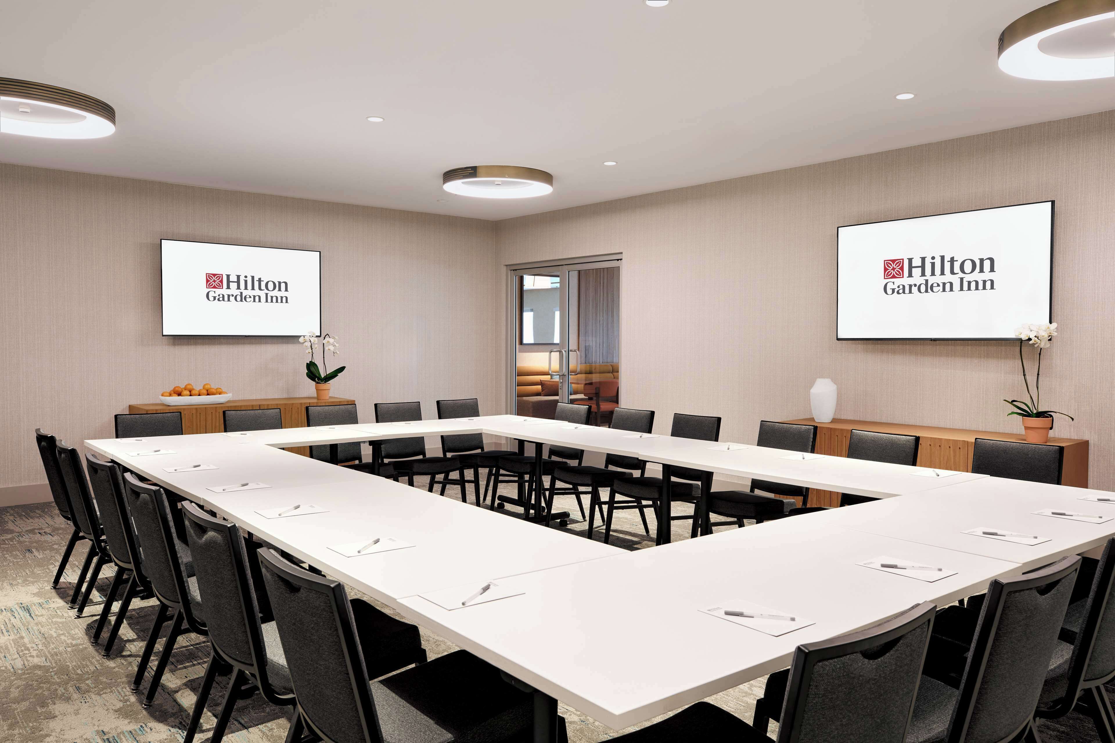 Un salón de conferencias con una larga mesa, sillas y dos pantallas mostrando el logotipo de Hilton Garden Inn.