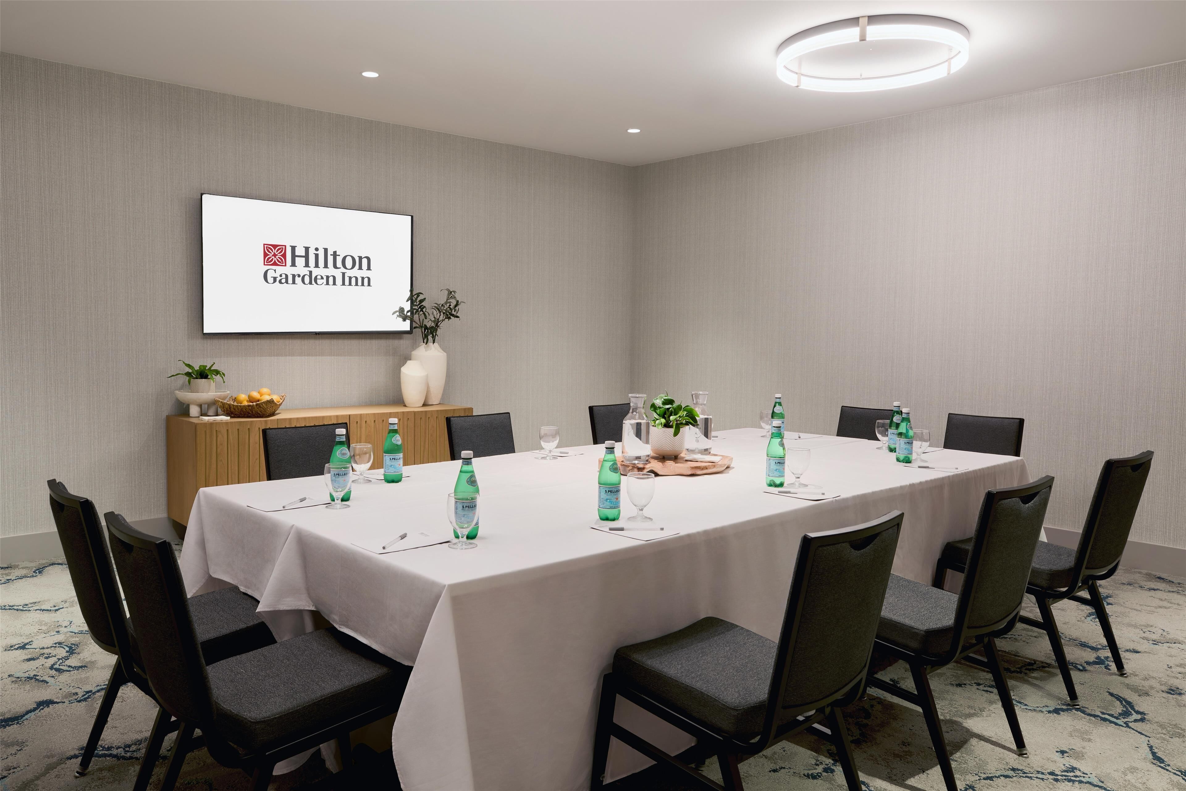 Une salle de conférence avec une longue table, des chaises, des bouteilles d’eau et une enseigne « Hilton Garden Inn » au mur.