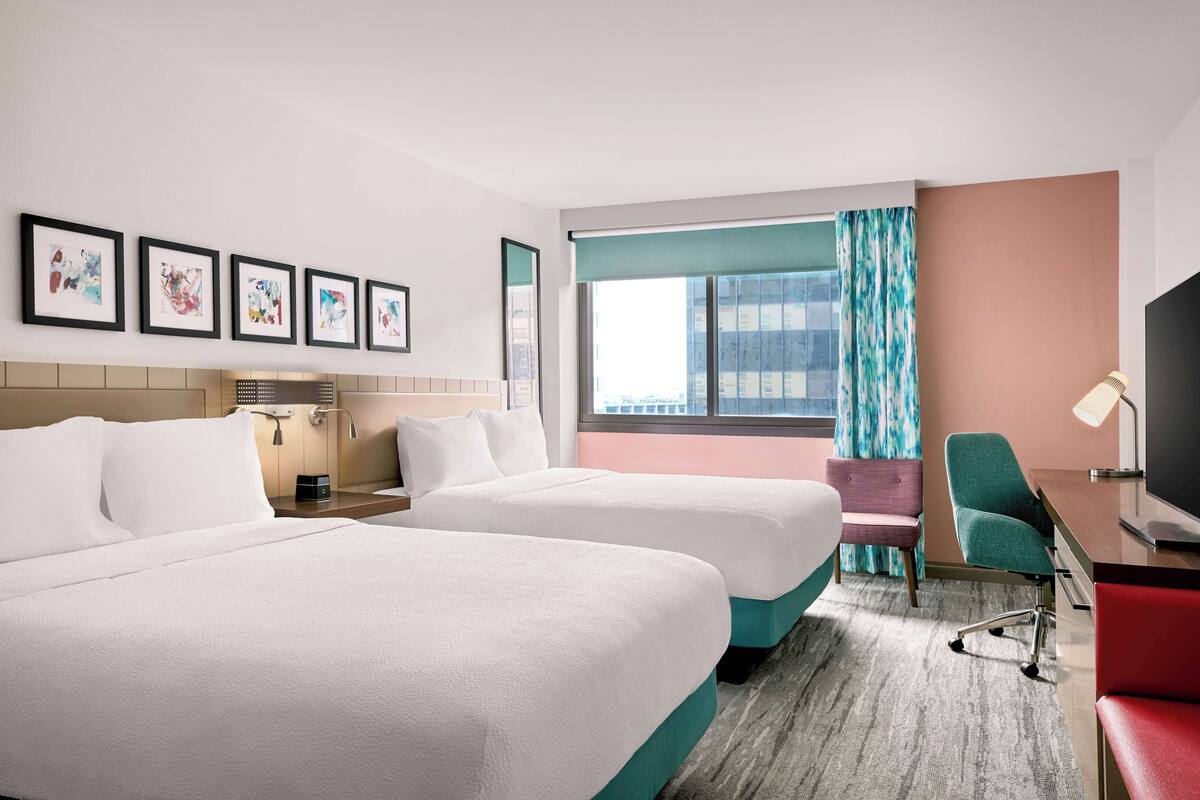 Une chambre d’hôtel avec deux lits, un bureau, une chaise, une télévision et des œuvres d’art accrochées au mur.