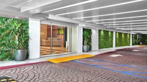 Entrada moderna de hotel con puerta de vidrio, plantas en macetas y piso de ladrillo estampado.