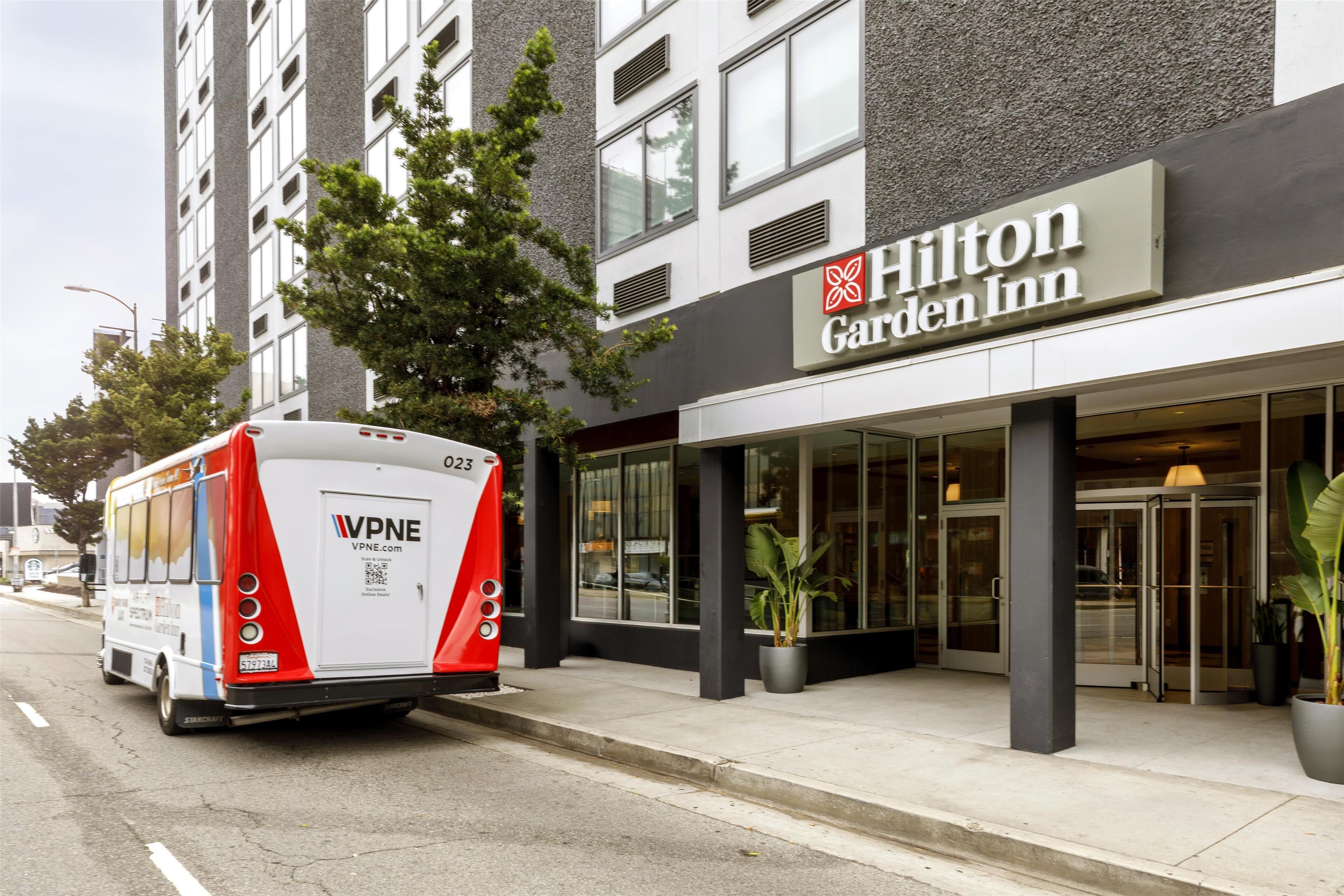 Ein modernes Gebäude mit einem Hilton Garden Inn Schild, einem Bus vor der Fassade und einem großen Kübelpflanzenarrangement in der Nähe des Eingangs.
