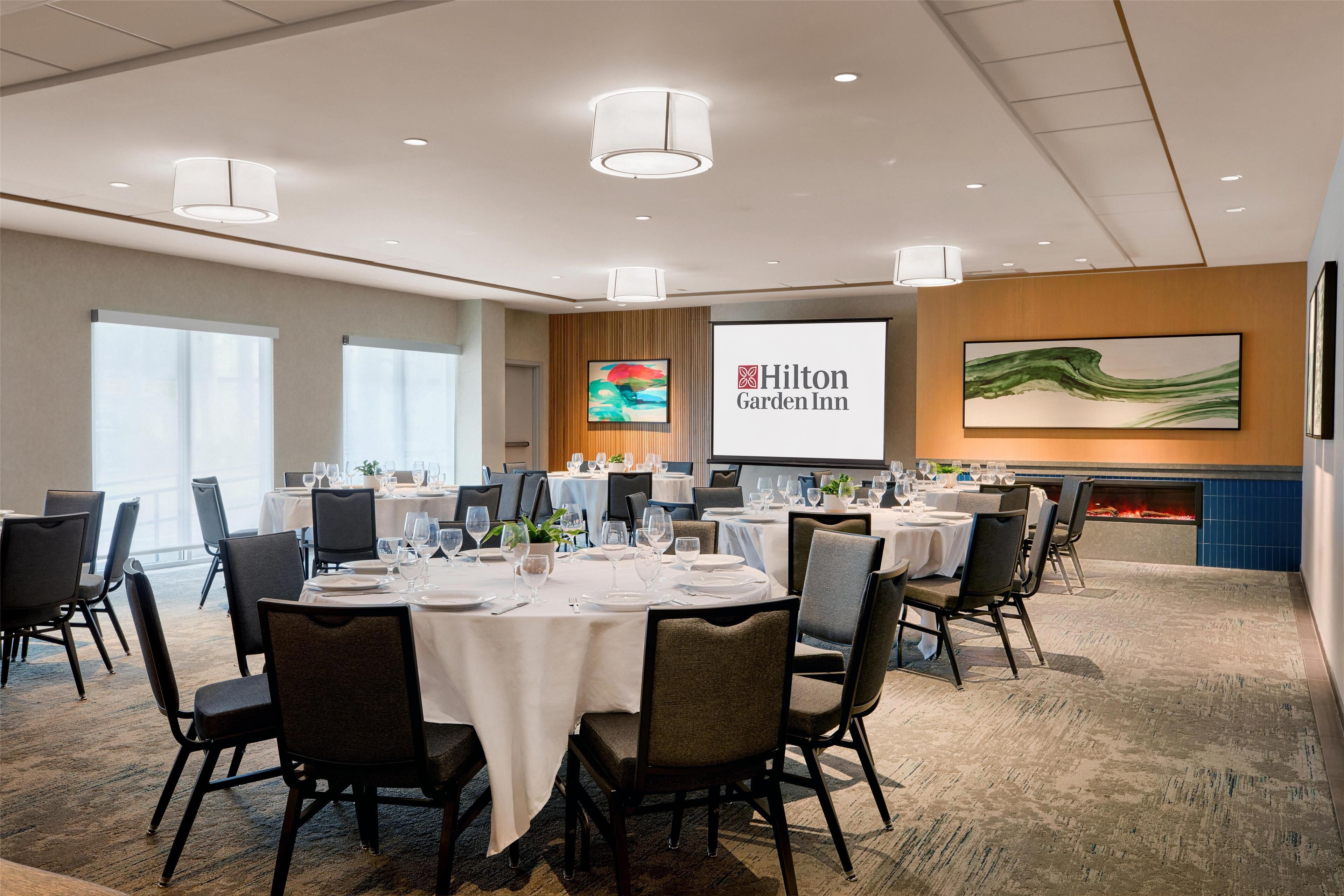 Ein Konferenzraum mit runden Tischen, Stühlen und einem großen Bildschirm, der das Logo des Hilton Garden Inn zeigt.