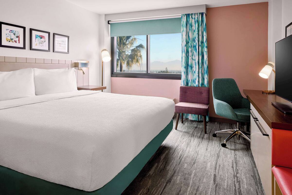 Une chambre d’hôtel équipée d’un lit, d’un bureau, d’une chaise, d’une télévision et d’une fenêtre donnant sur des palmiers.