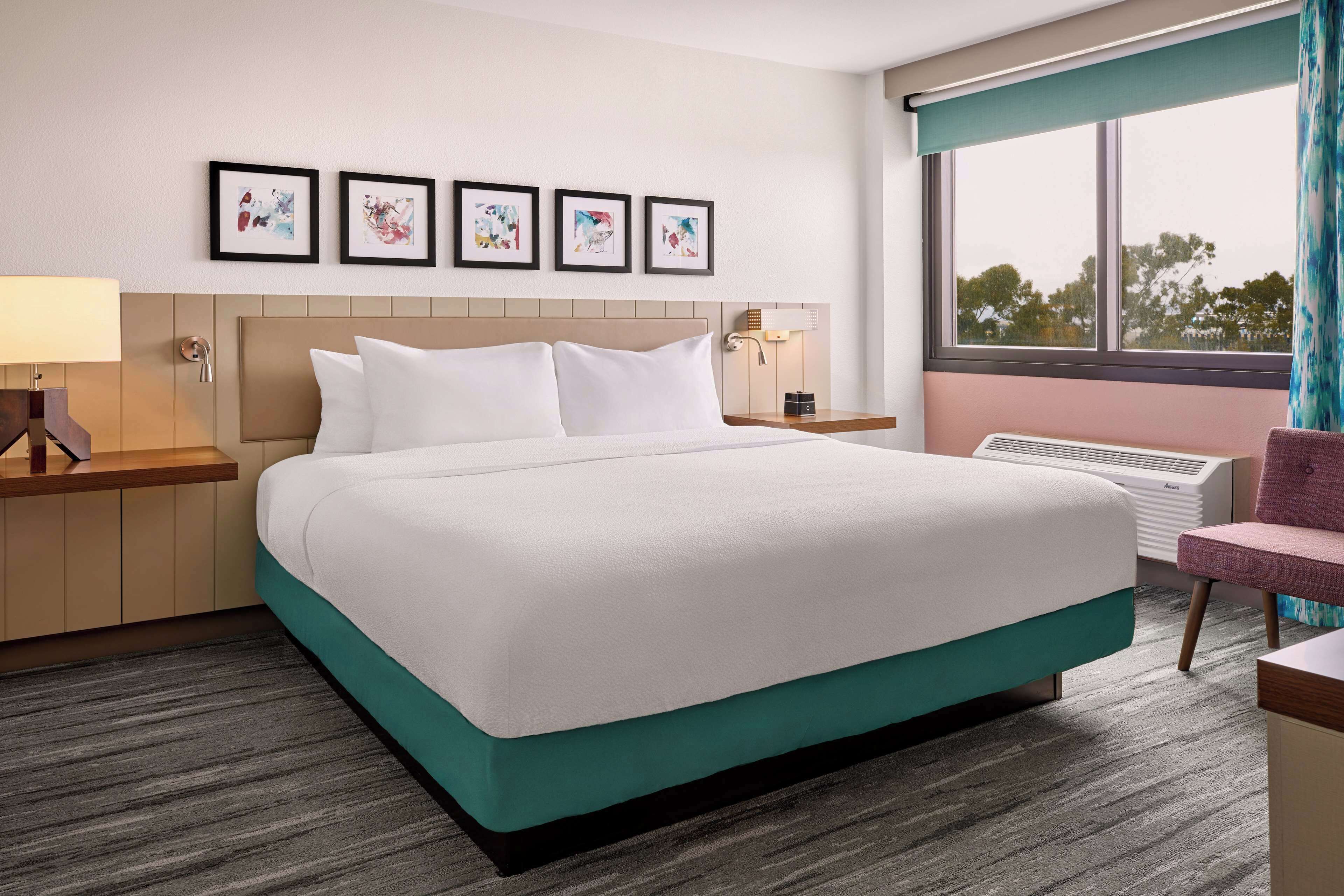A hotel room with a large bed, a desk, a chair, and a window with a view of trees.