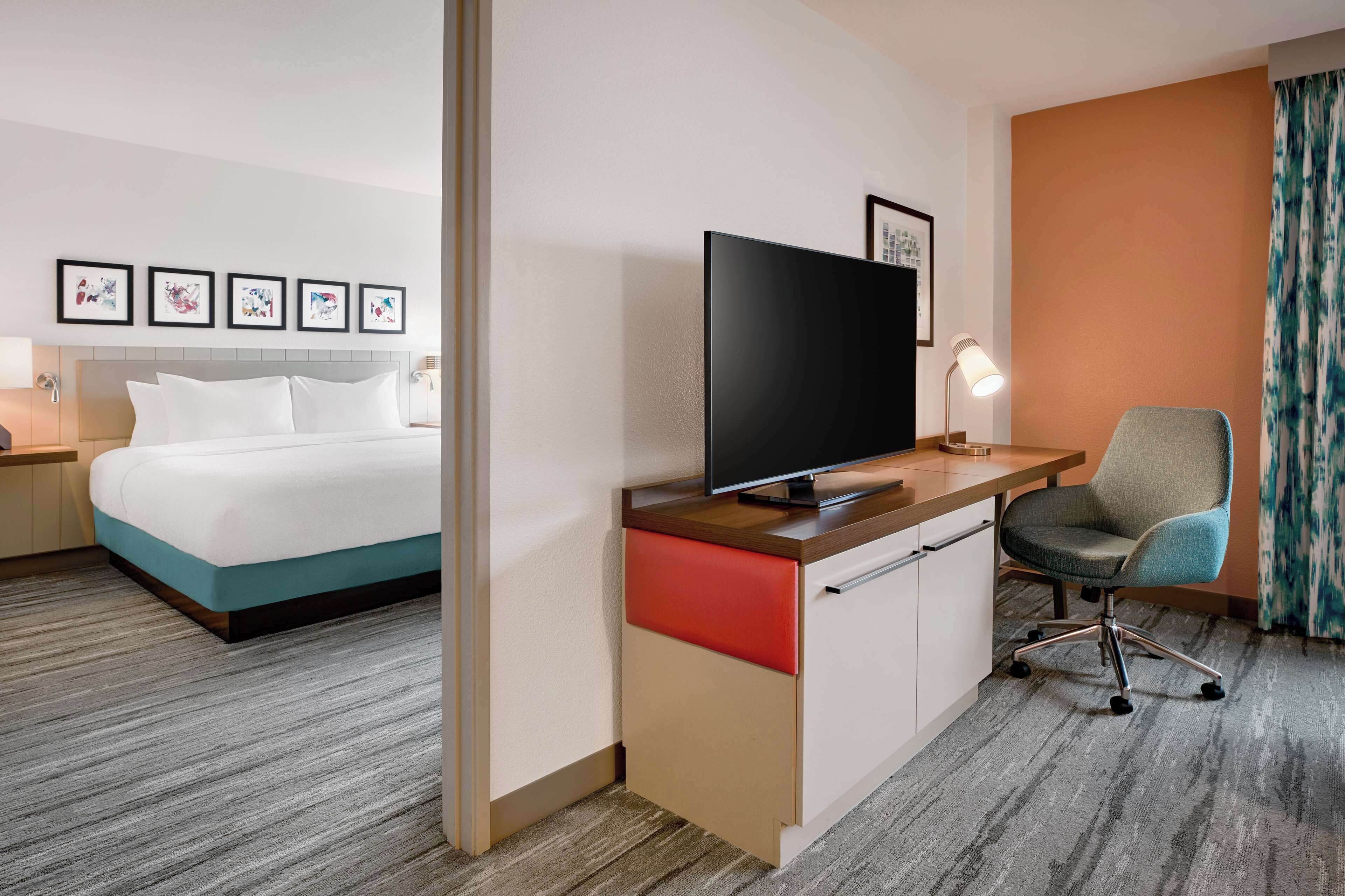 Una habitación de hotel con una cama, un escritorio con televisor, una silla, una lámpara y cuadros en la pared.