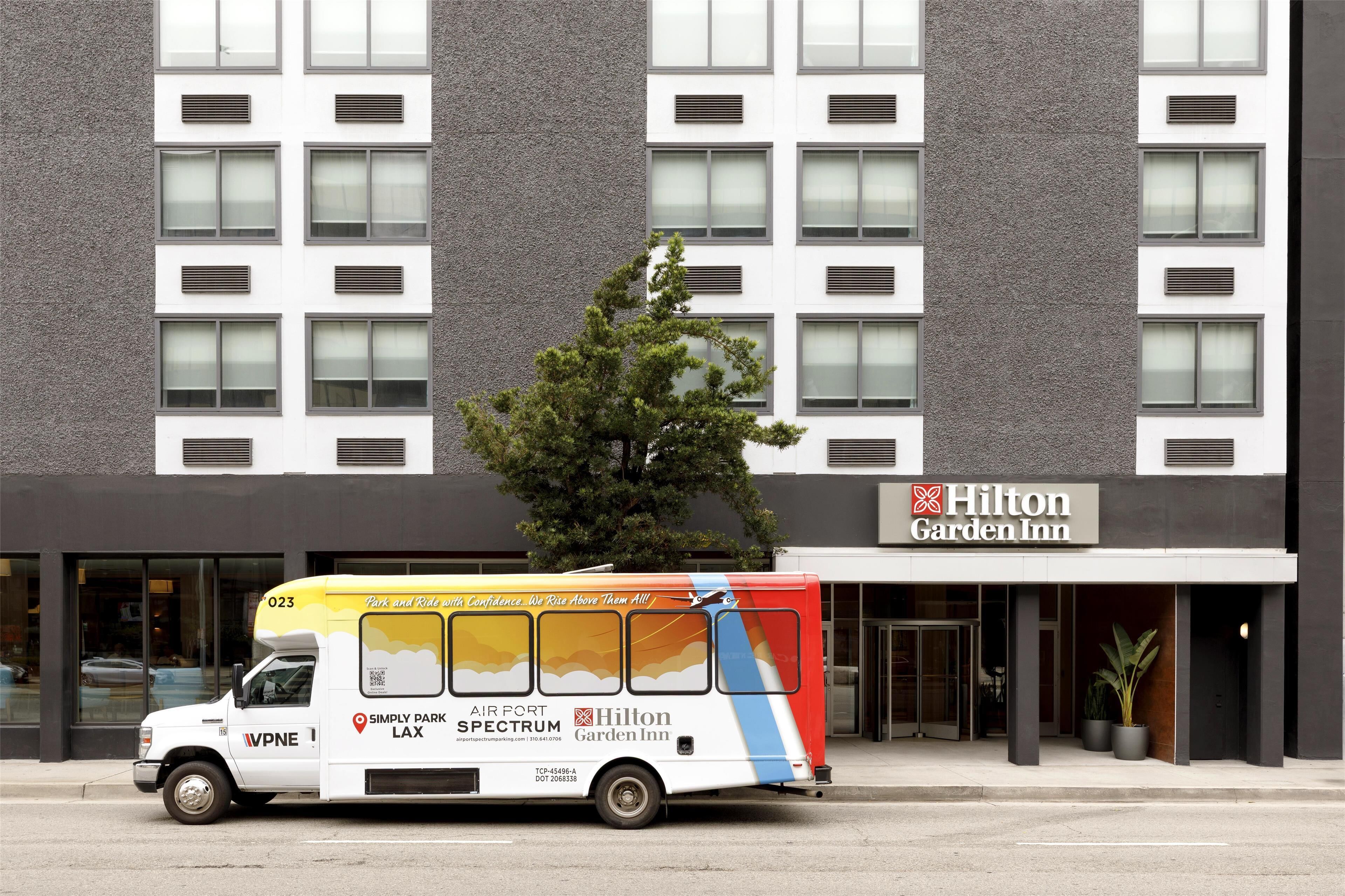 Un camion de livraison blanc et jaune est garé devant un bâtiment de l’hôtel Hilton Garden Inn, avec un grand arbre devant.