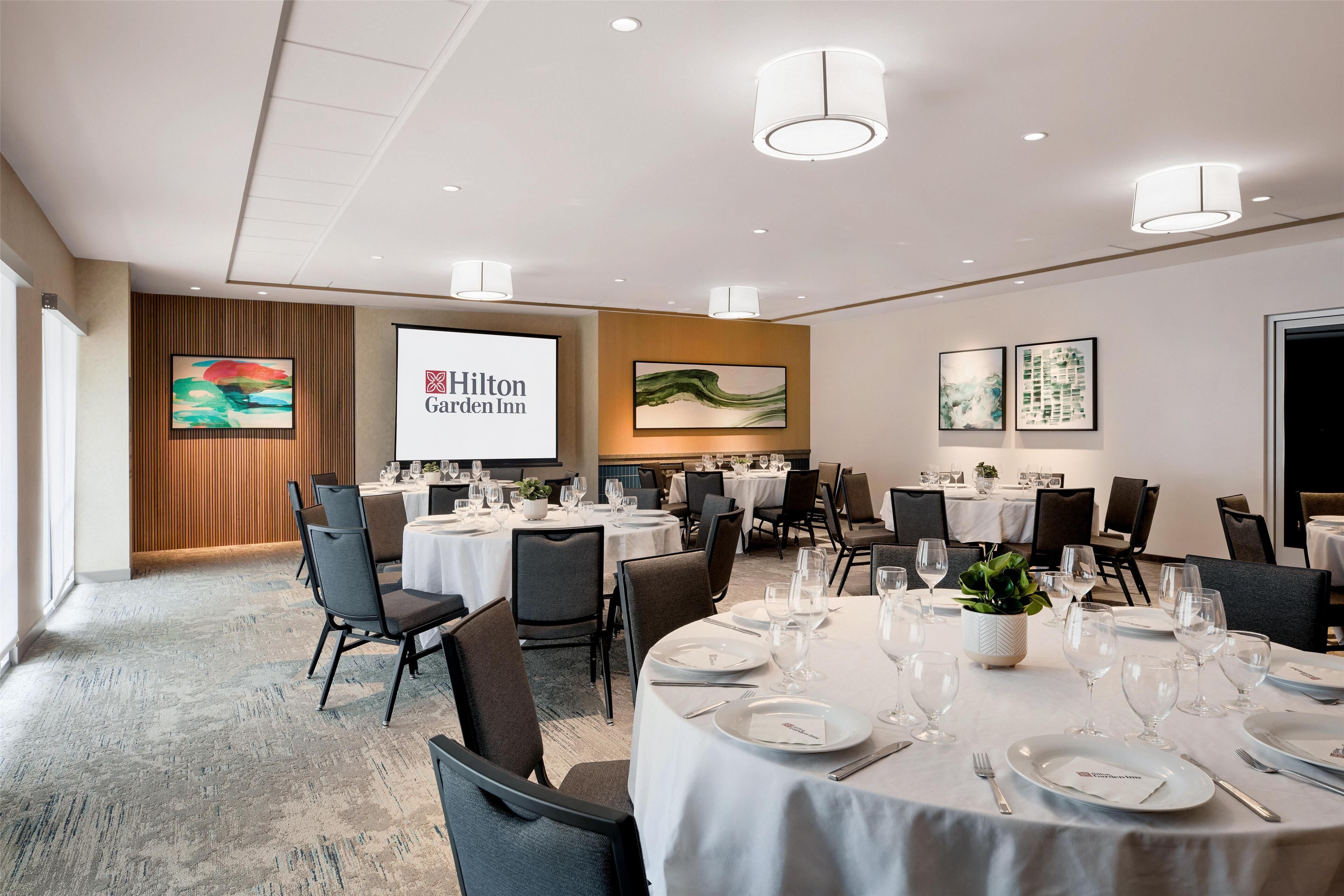 Uma sala de conferências bem iluminada com mesas redondas preparadas para uma reunião, uma tela grande exibindo o logotipo do Hilton Garden Inn e obras de arte emolduradas nas paredes.