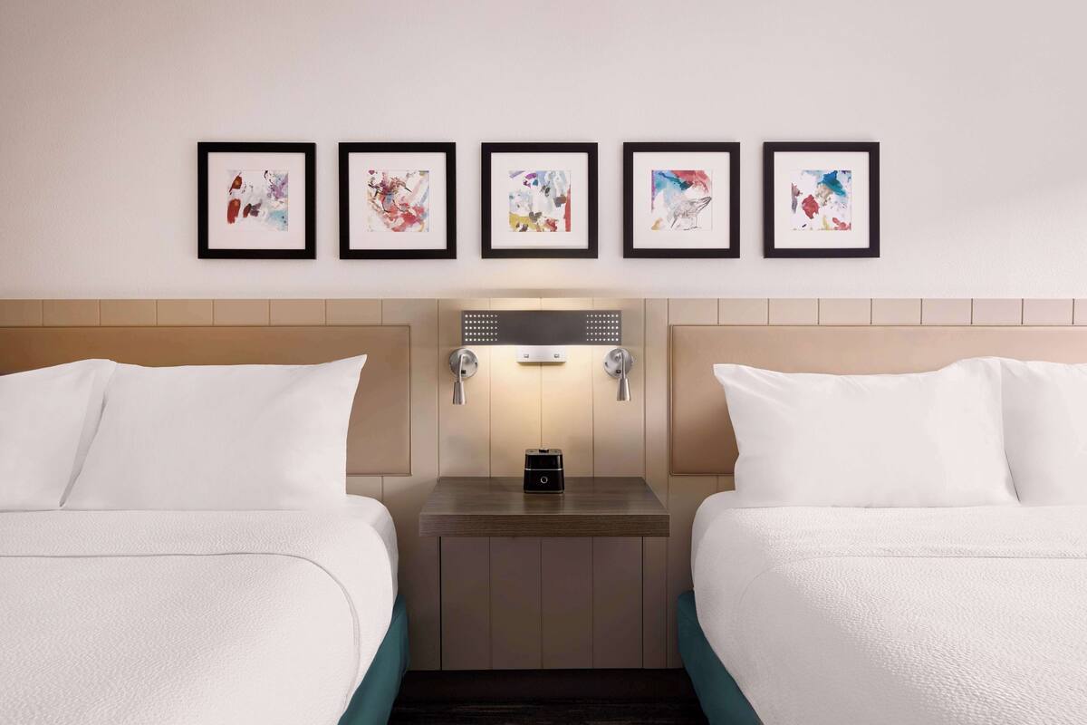 Une chambre d’hôtel avec deux lits, une table de chevet avec une lampe et cinq œuvres d’art encadrées accrochées au mur.