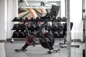 Uma bicicleta ergométrica numa academia, com um suporte de halteres ao fundo.