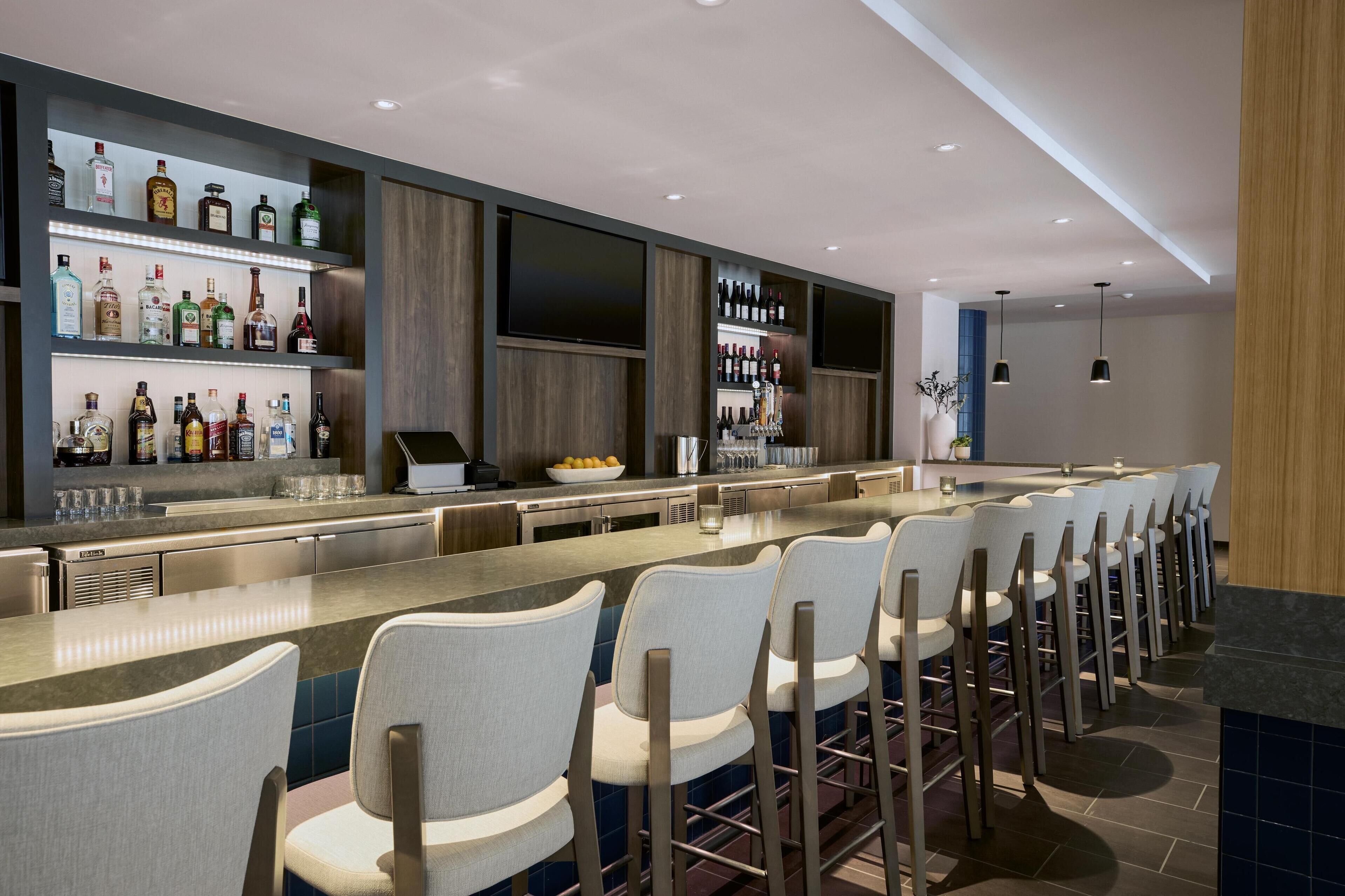A modern bar area with a long counter, multiple seating options, and a variety of liquor bottles on shelves.