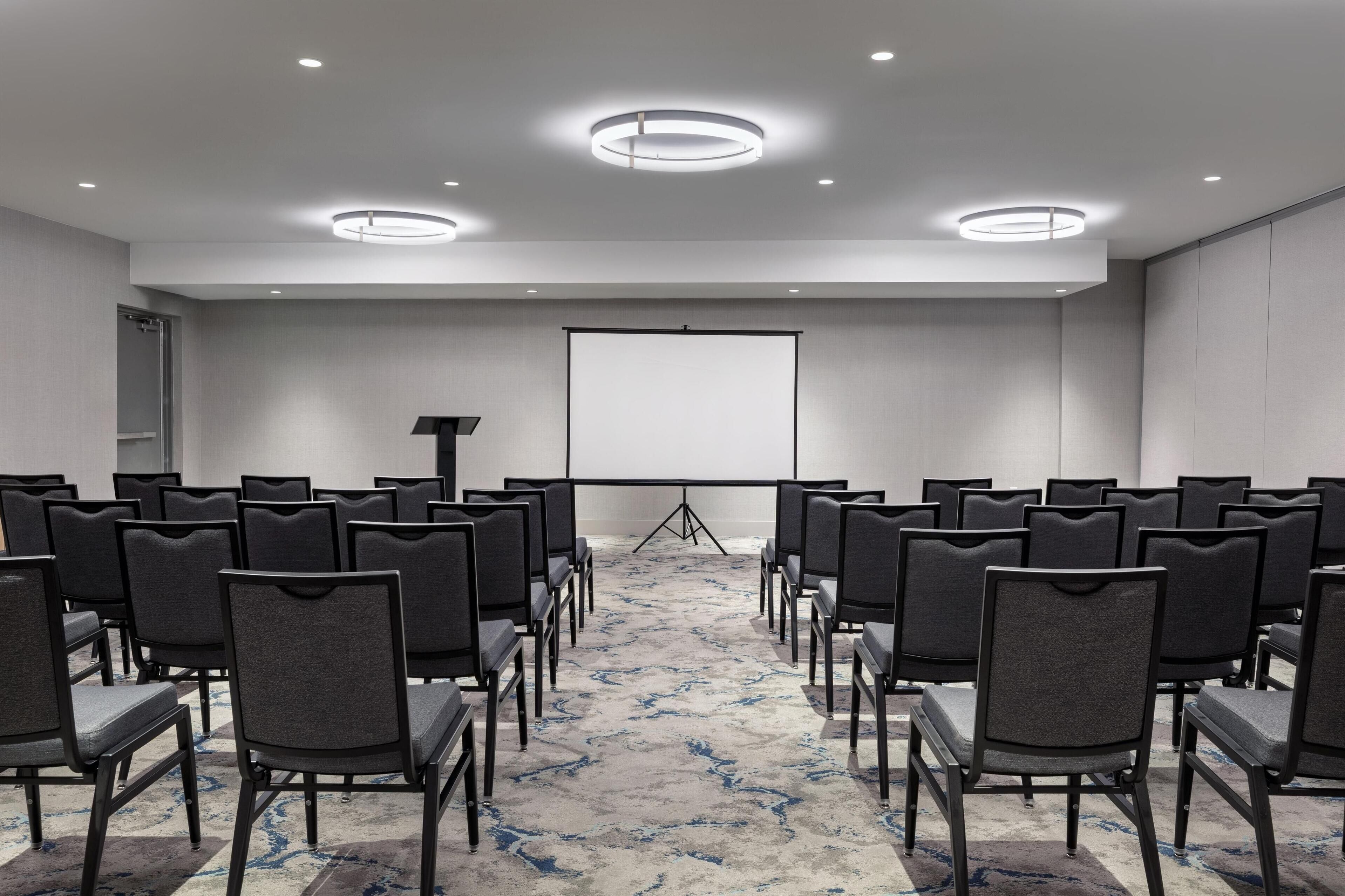 A conference room with rows of chairs facing a projection screen, a podium, and a carpeted floor.