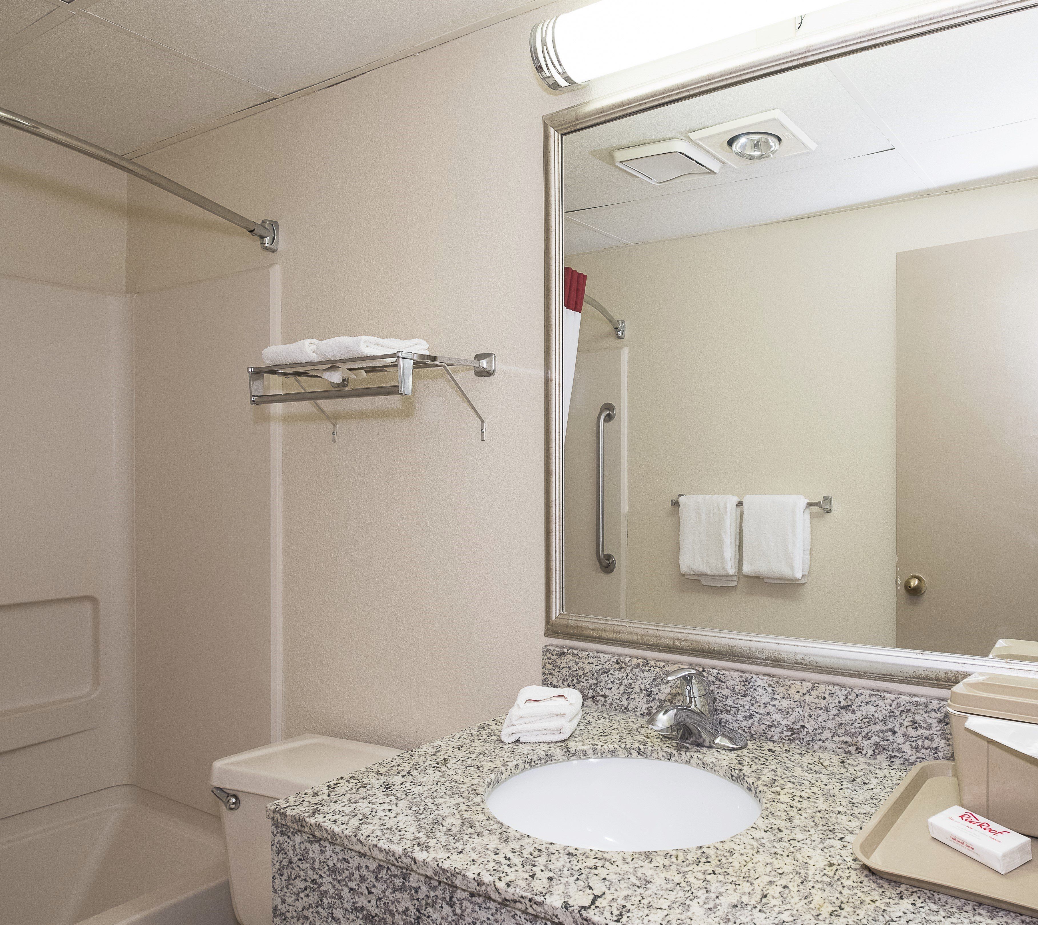 Un baño con lavamanos de encimera de granito, inodoro, ducha con barra de apoyo y espejo que refleja un perchero y una puerta.