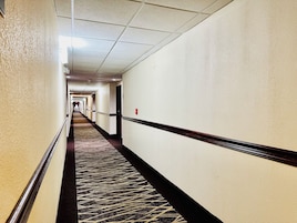 Corredor de hotel com piso de carpete, paredes brancas e portas que dão acesso aos quartos.
