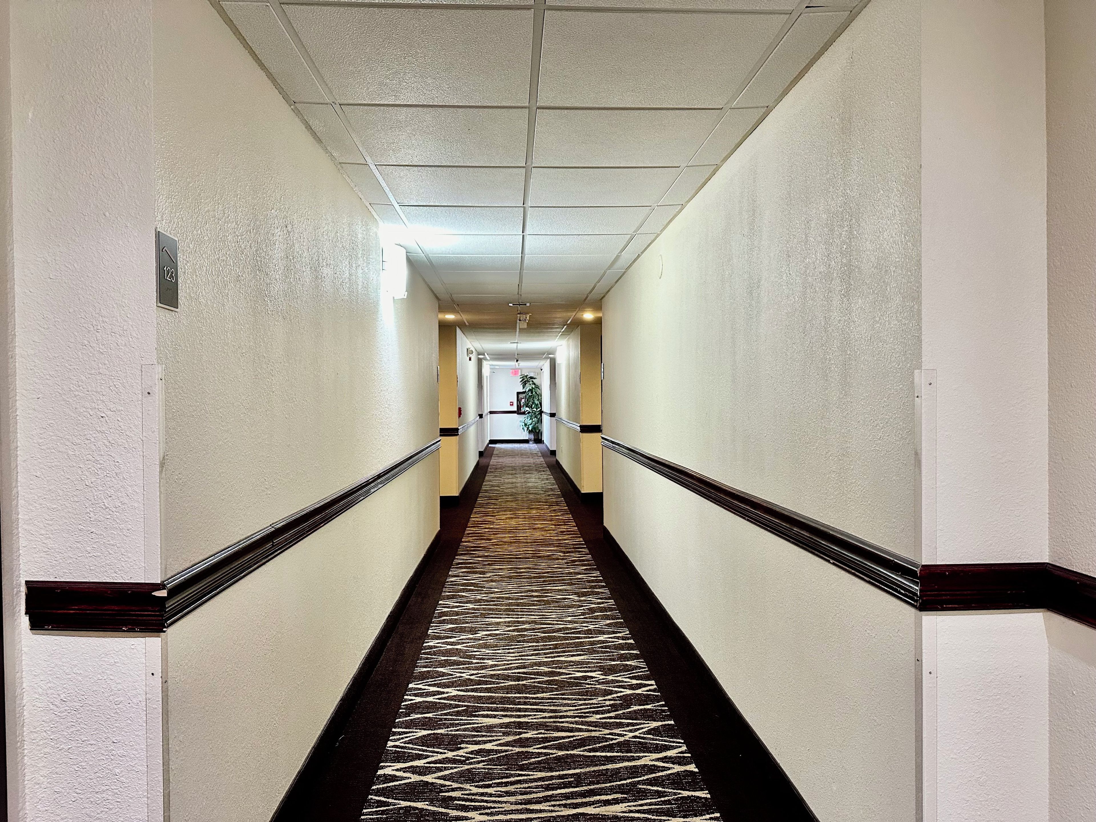A long, narrow hotel corridor with a patterned carpet, white walls, and a ceiling with recessed lighting.
