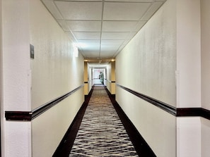 Un largo y estrecho pasillo de hotel con alfombra estampada, paredes blancas e iluminación empotrada en el techo.