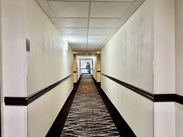Un largo y estrecho pasillo de hotel con alfombra estampada, paredes blancas e iluminación empotrada en el techo.