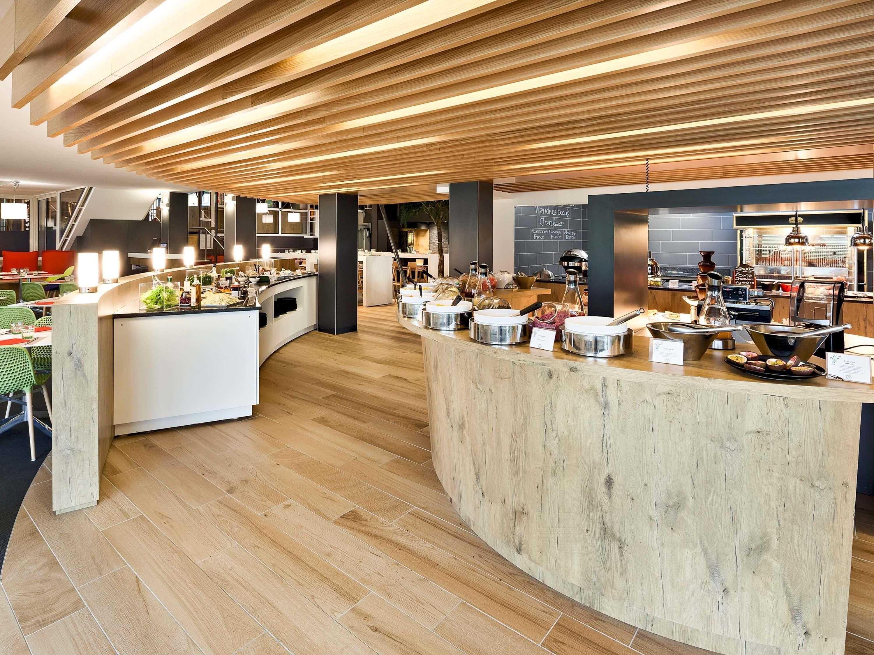 A modern cafe with a wooden ceiling, a central serving counter, and a seating area with tables and chairs.