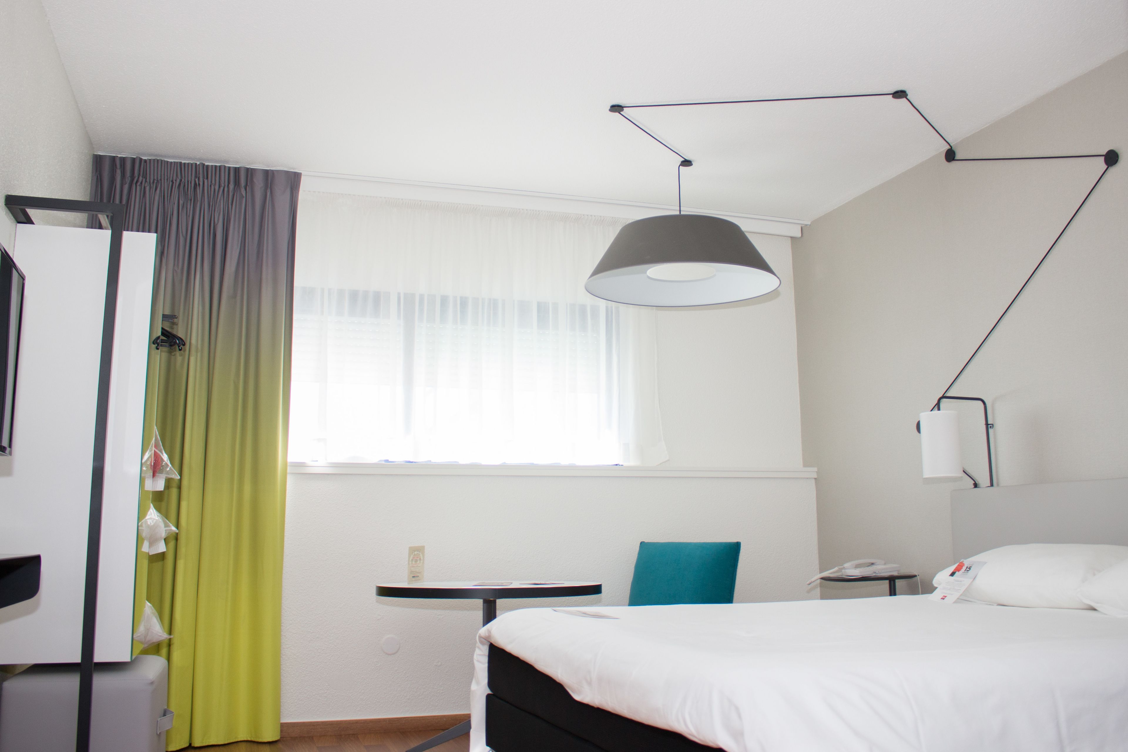 A hotel room with a bed, a desk, a chair, a lamp, and a window with curtains.