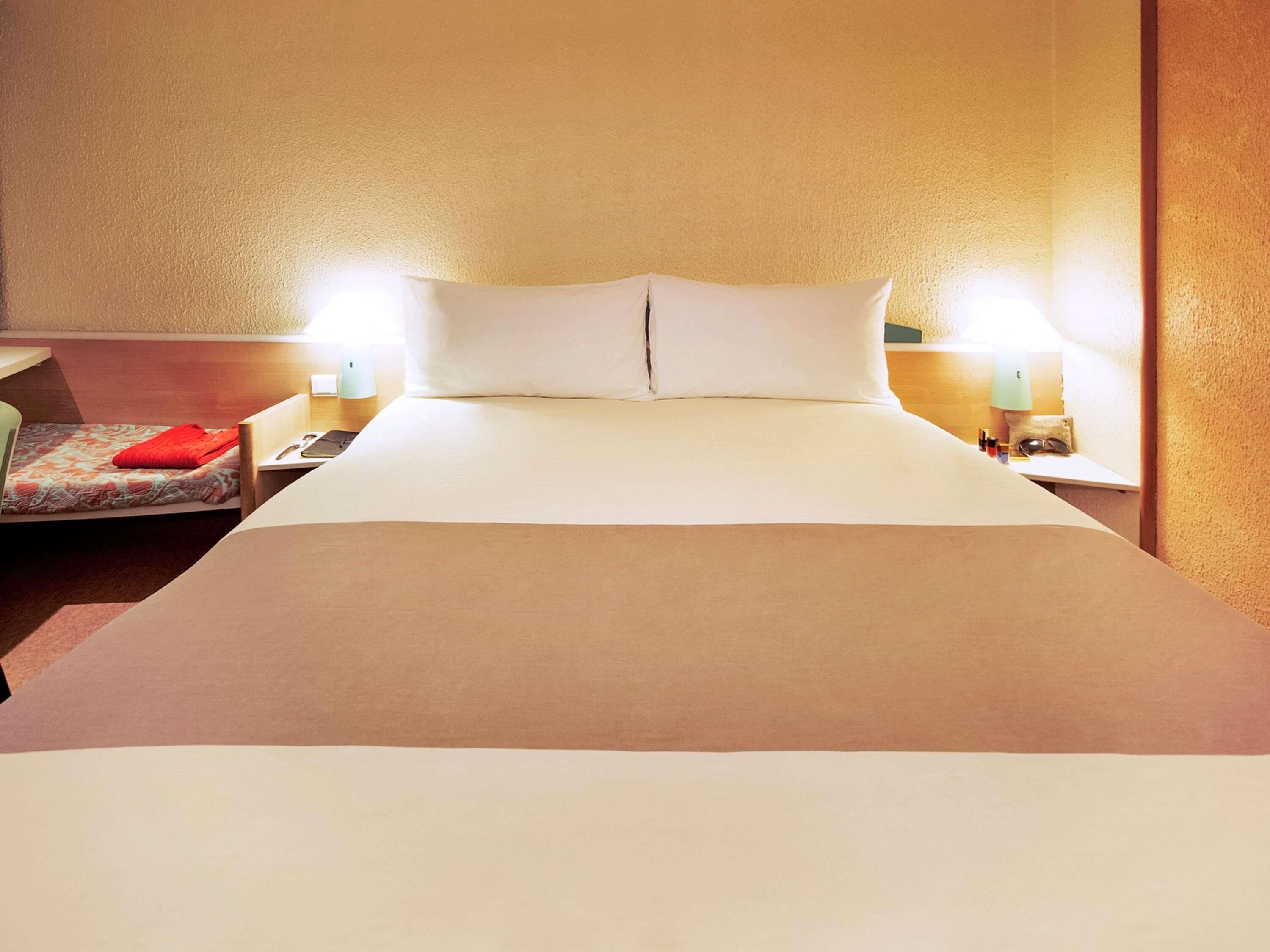 Una habitación de hotel con una cama grande, dos mesitas de noche, una silla y una lámpara.