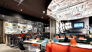 A modern restaurant with a bar area, seating with orange cushions, and a large screen displaying content.