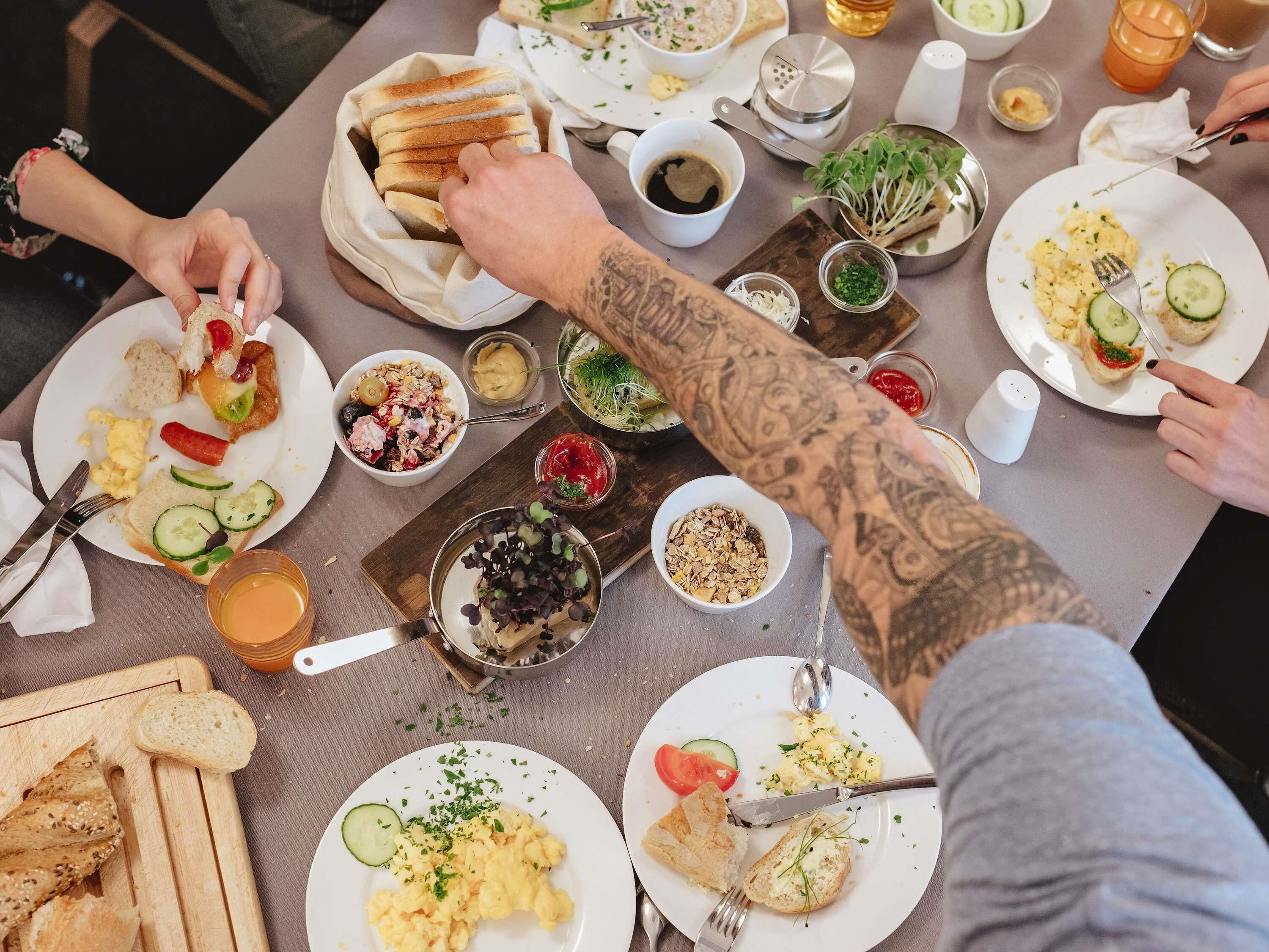 Ein Esstisch mit verschiedenen Frühstücksangeboten, Besteck und einer Tätowierten Person.