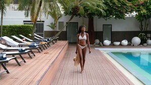 Una terraza de madera con sillas de descanso y zona junto a la piscina, palmeras y un edificio al fondo.