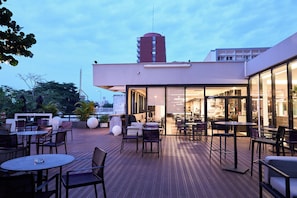An outdoor patio area with tables and chairs, a building entrance, and a tall structure in the background.