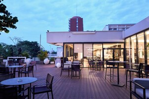 Un espacio exterior con patio, mesas y sillas, la entrada de un edificio y una estructura alta al fondo.