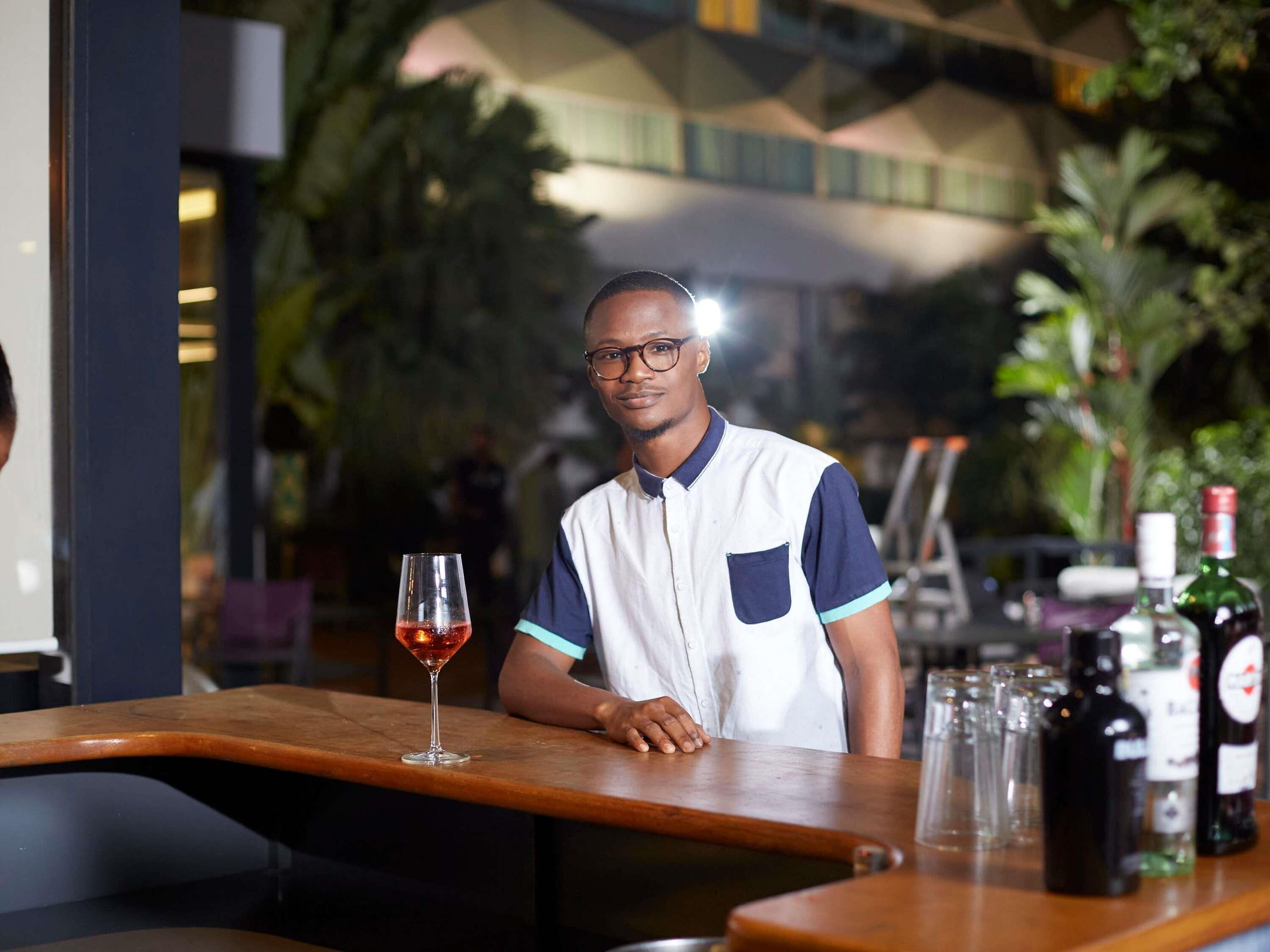 Un barman derrière le comptoir d’un bar, avec une boisson dans un verre, des bouteilles et d’autres verres à proximité.